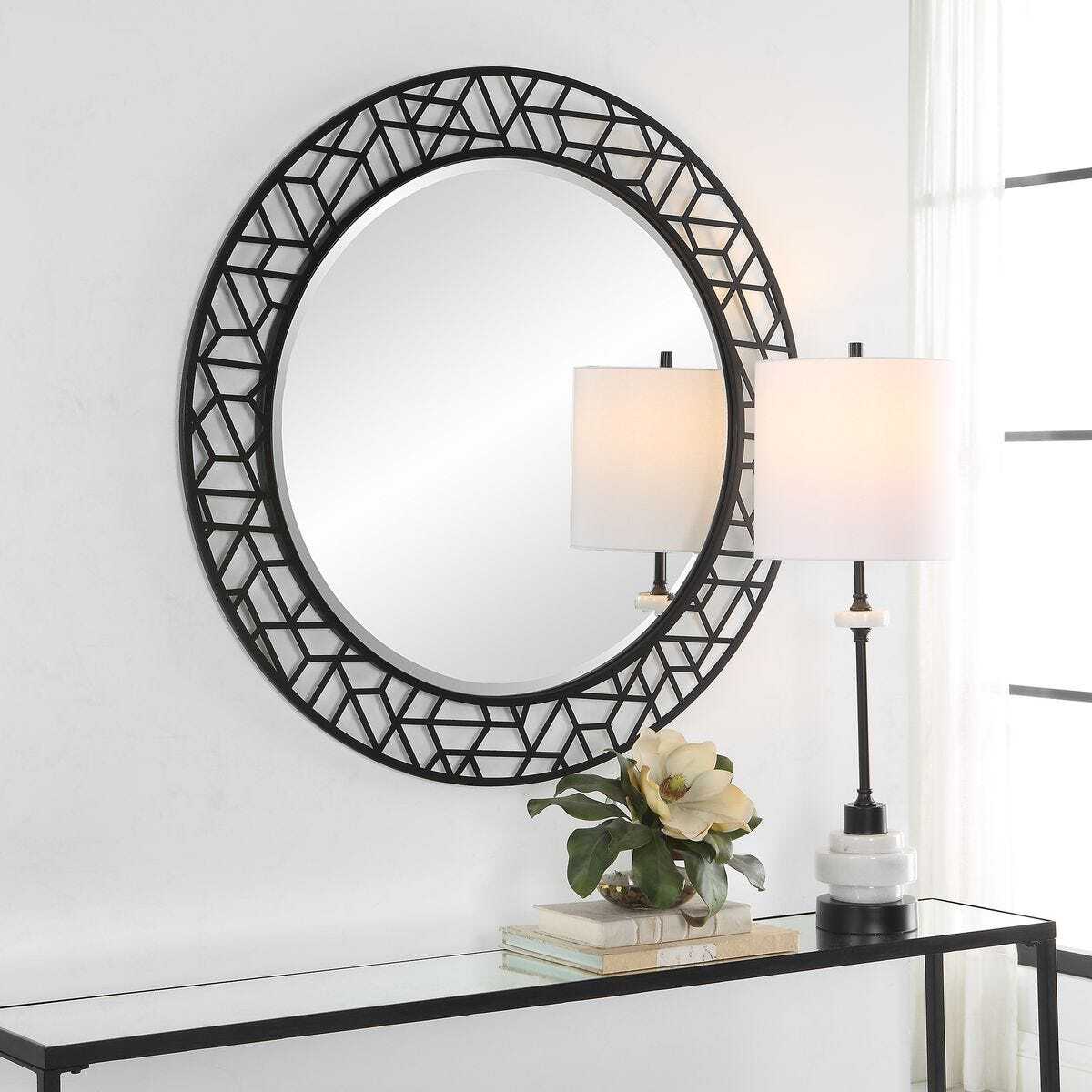 Round wall mirror with geometric black frame on a white wall, next to a table with decor items.