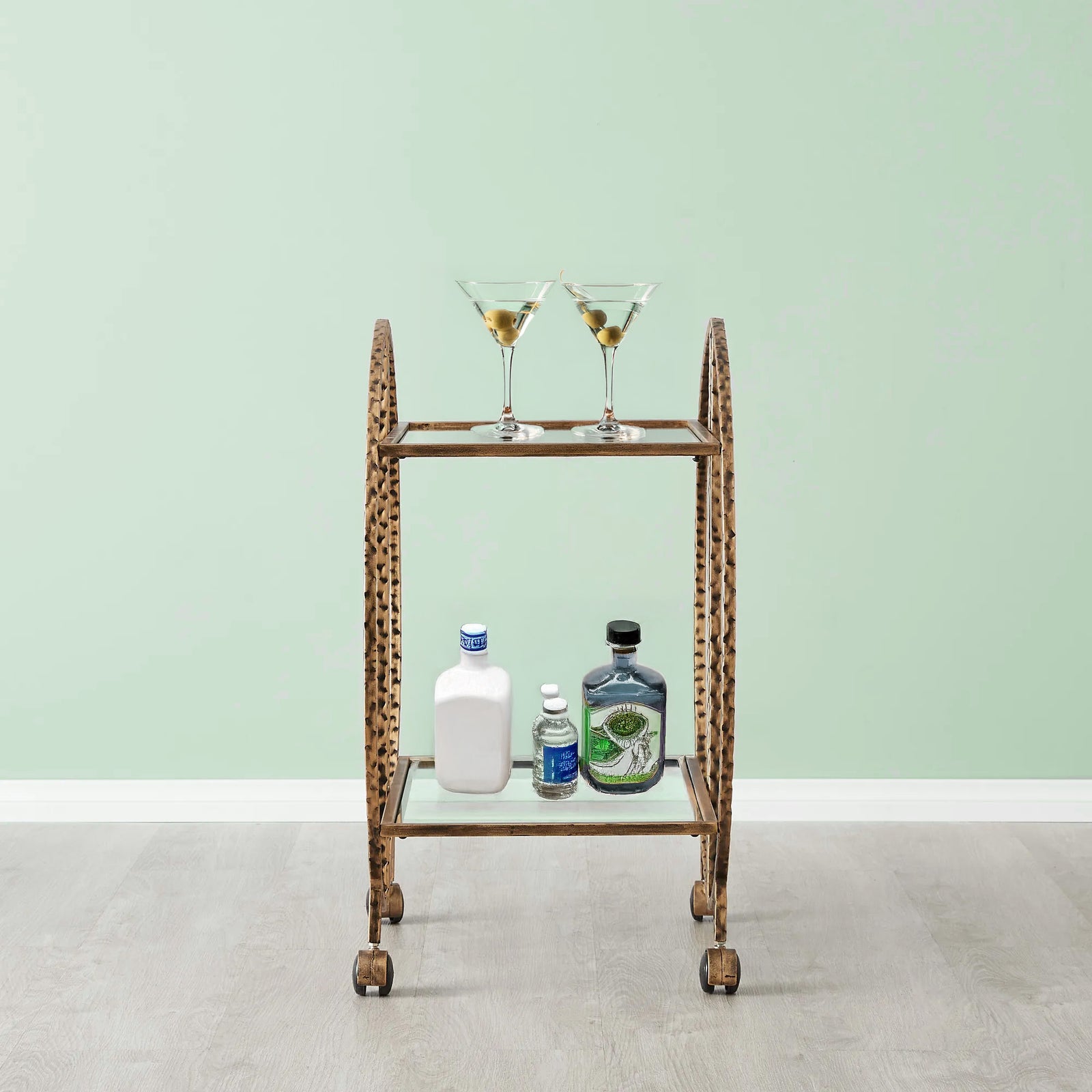 An old bart cart with hammered metal in the color antique gold. The back wall is a sea-foam green. Bottles of liquor are on the bottom shelf of the bar cart.