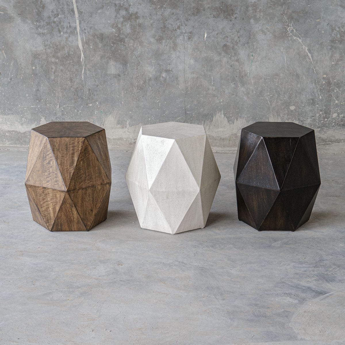 Three geometrically shaped stools in wood, concrete, and dark material against a gray concrete wall.