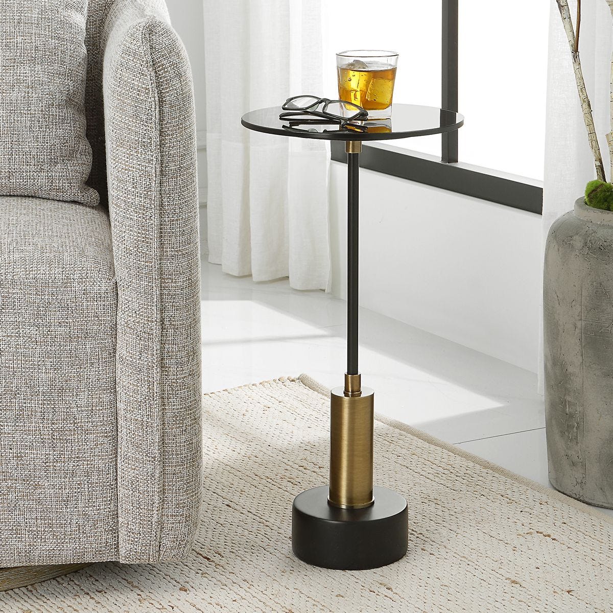 Small round side table with black base and gold cylindrical accent next to a gray sofa.