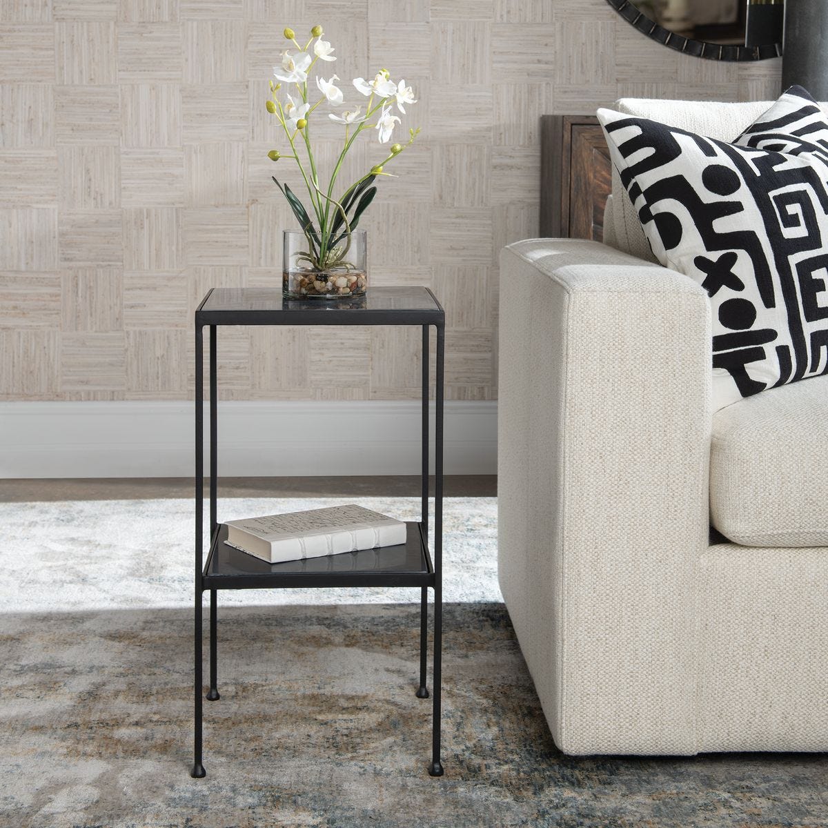 Black side table with a plant and book next to a beige sofa with patterned cushions.