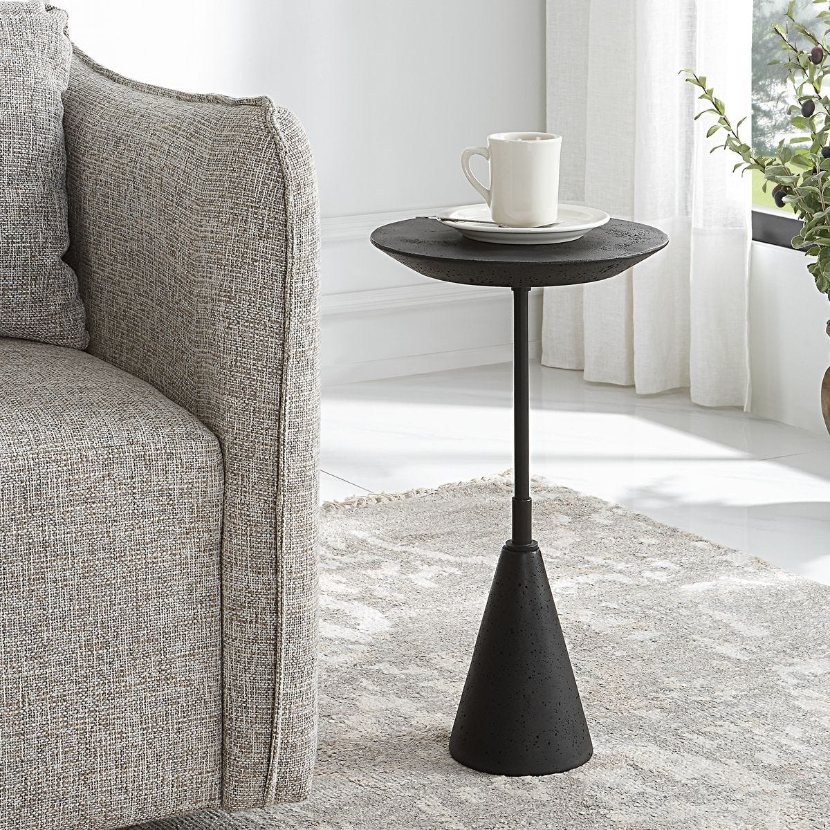 Black side table with a white mug and saucer next to a gray sofa in a living room.