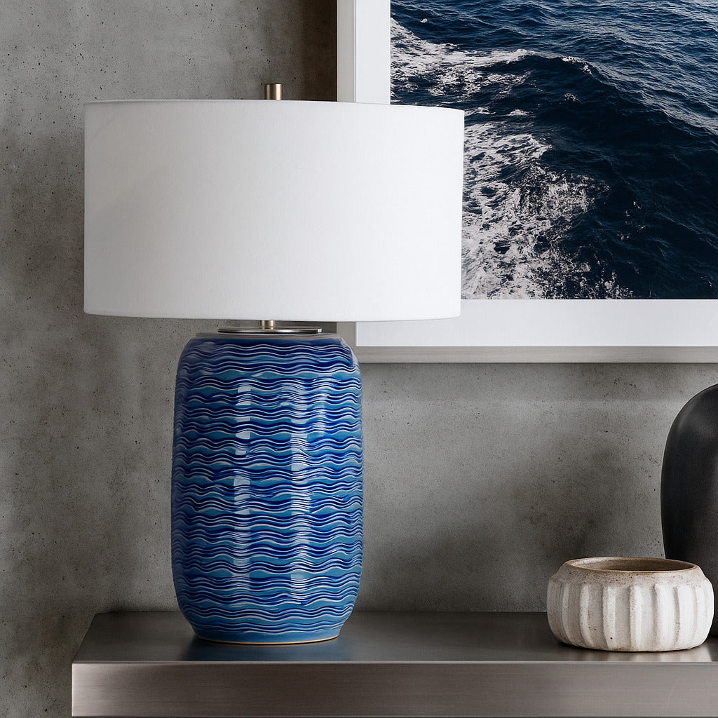 Blue textured table lamp with white shade on a shelf against a concrete wall with ocean art.