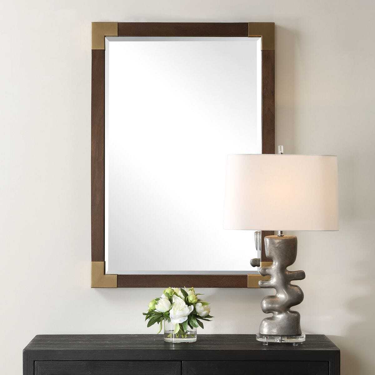 Decorative mirror with wooden frame above a table with a lamp and floral arrangement.