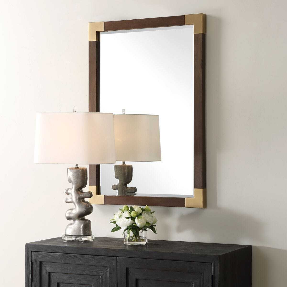 Decorative mirror with gold frame above a dark wooden console table with a lamp and flowers.