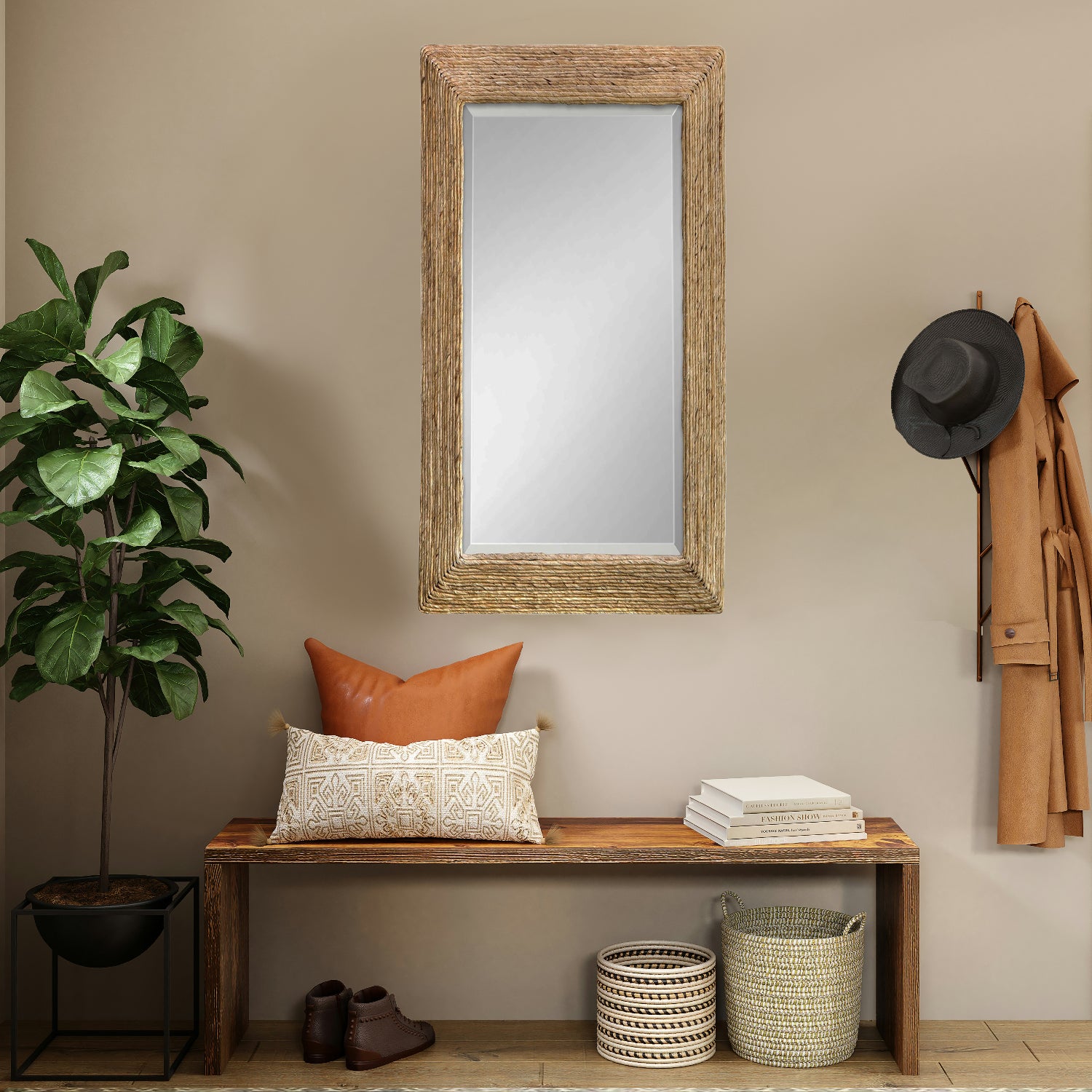 Large woven coastal mirror above bench with pillows and books on it. Hanging coat is by the front door and a fiddle fig plant in the corner with shoes and baskets underneath the bench.