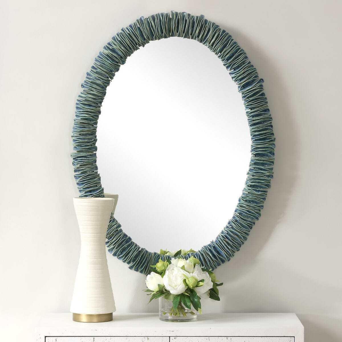 Round mirror with textured blue frame on a white surface with a vase and flowers.