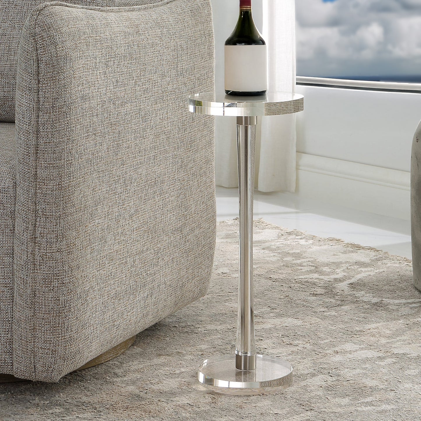 Round clear drink table with bottle of wine on top in a coastal condo with a gray rug and decorative chair.