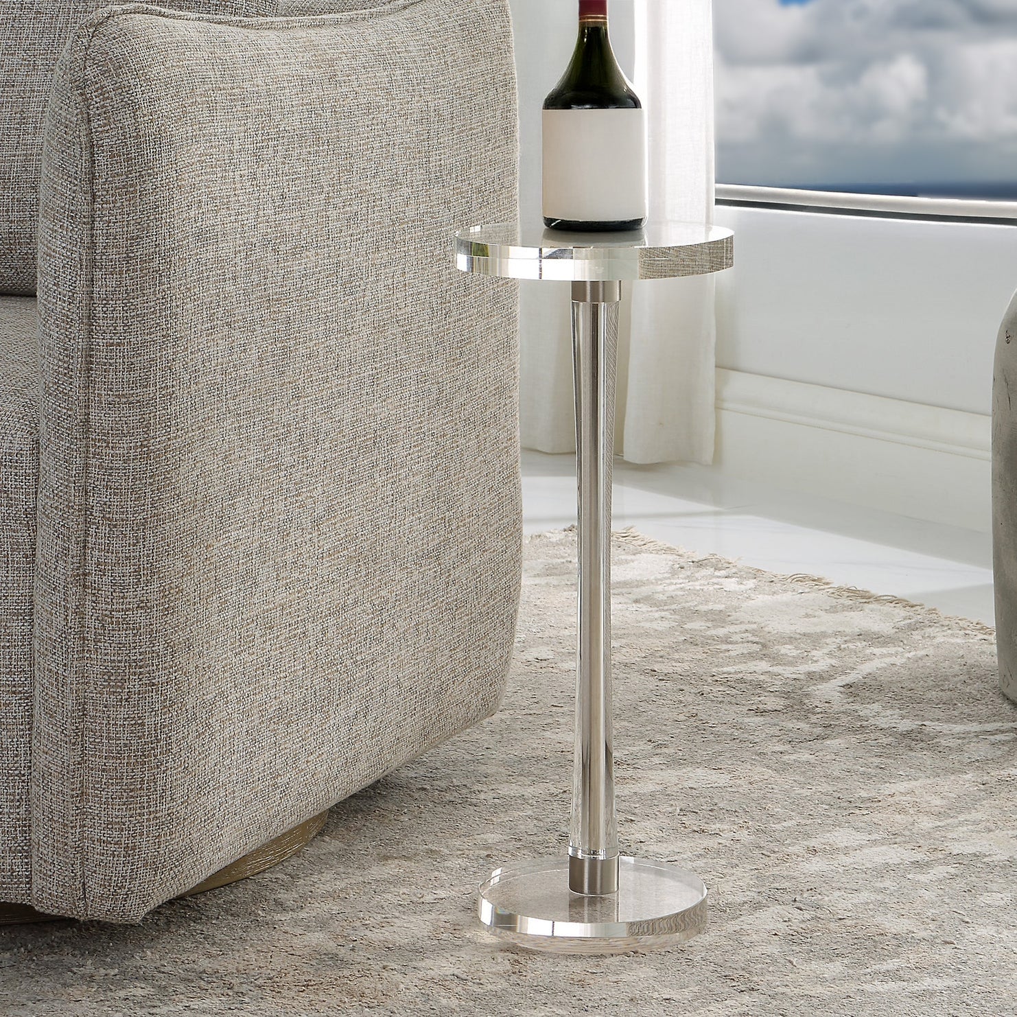 Round clear drink table with bottle of wine on top in a coastal condo with a gray rug and decorative chair.