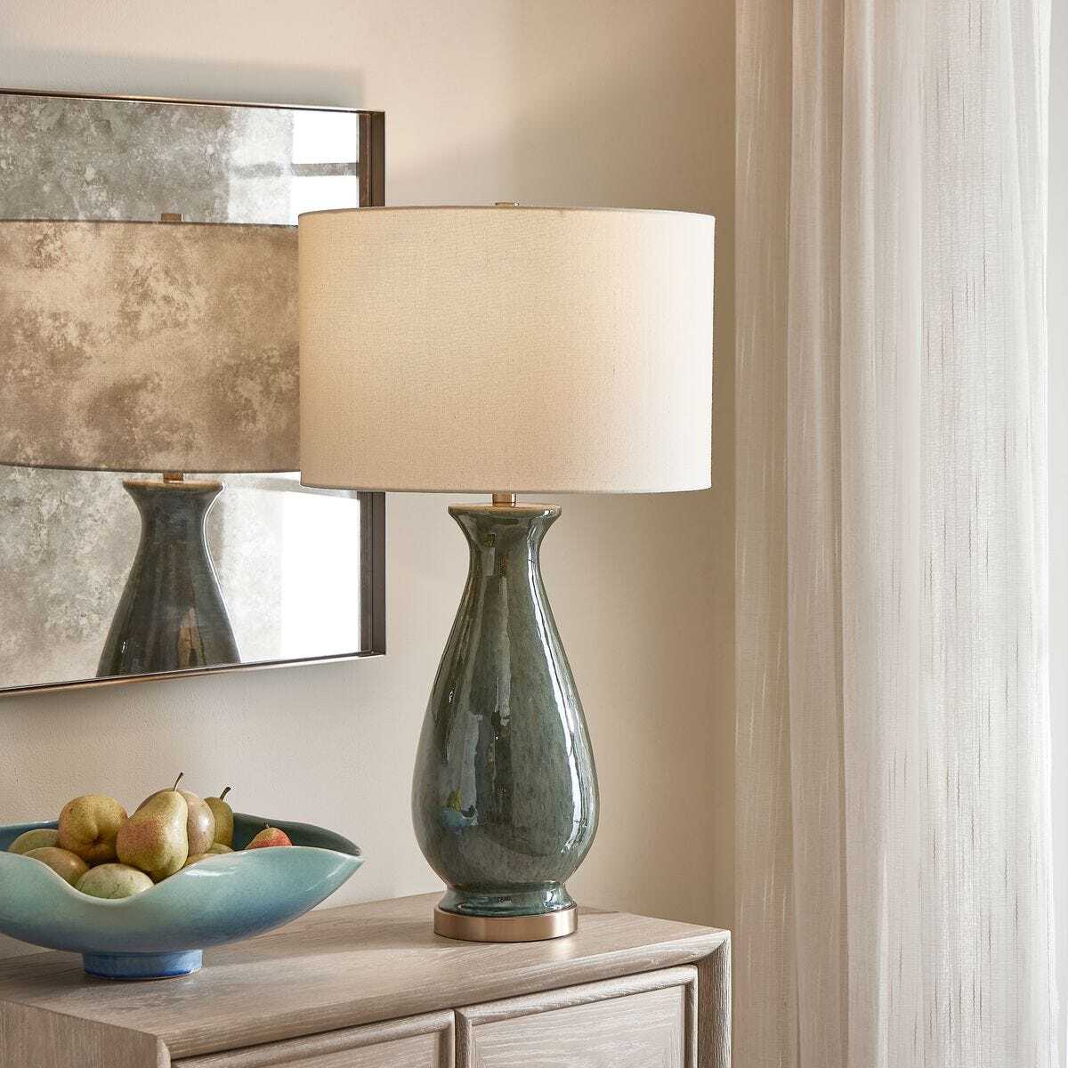 Green ceramic table lamp with beige lampshade on a wooden surface, with a decorative mirror and fruit bowl in the background.