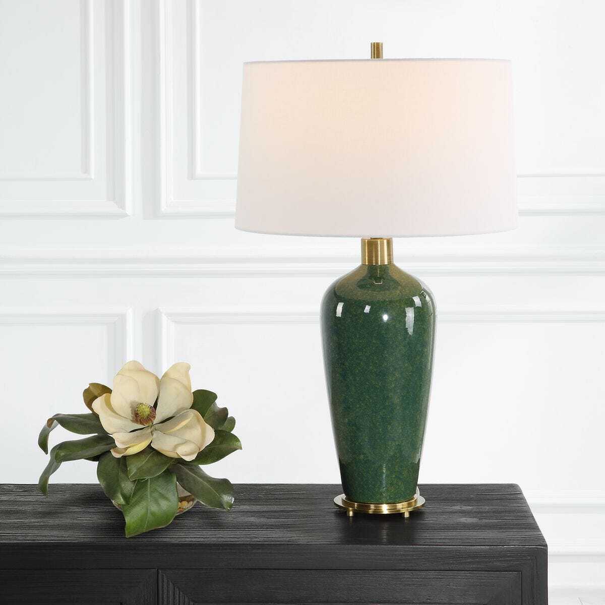 Green table lamp with white shade on a dark surface against a white paneled wall.