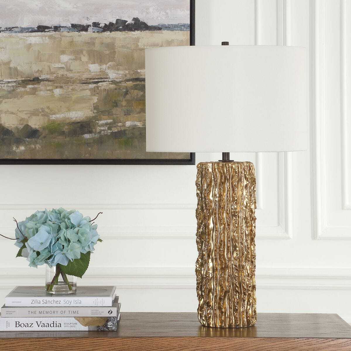 Decorative lamp with textured base on a shelf with books and flowers, against a white wall with framed artwork.