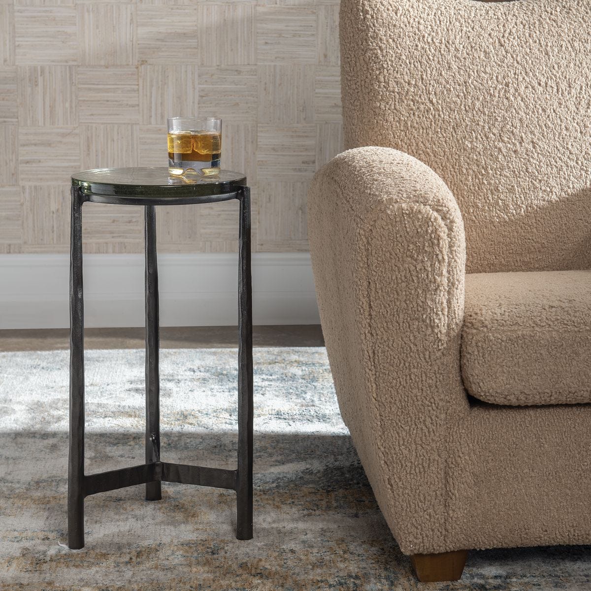 Small black side table with a glass of amber liquid next to a beige sofa against a patterned wall.