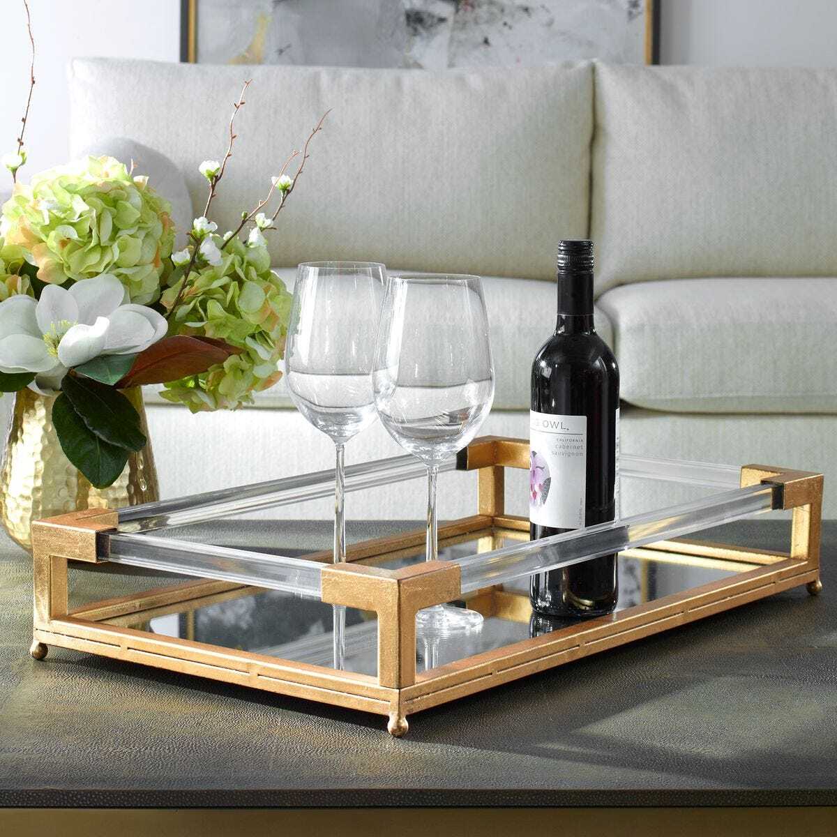 Glass tray with wine glasses and a bottle on a coffee table, with a couch and decorative items in the background.