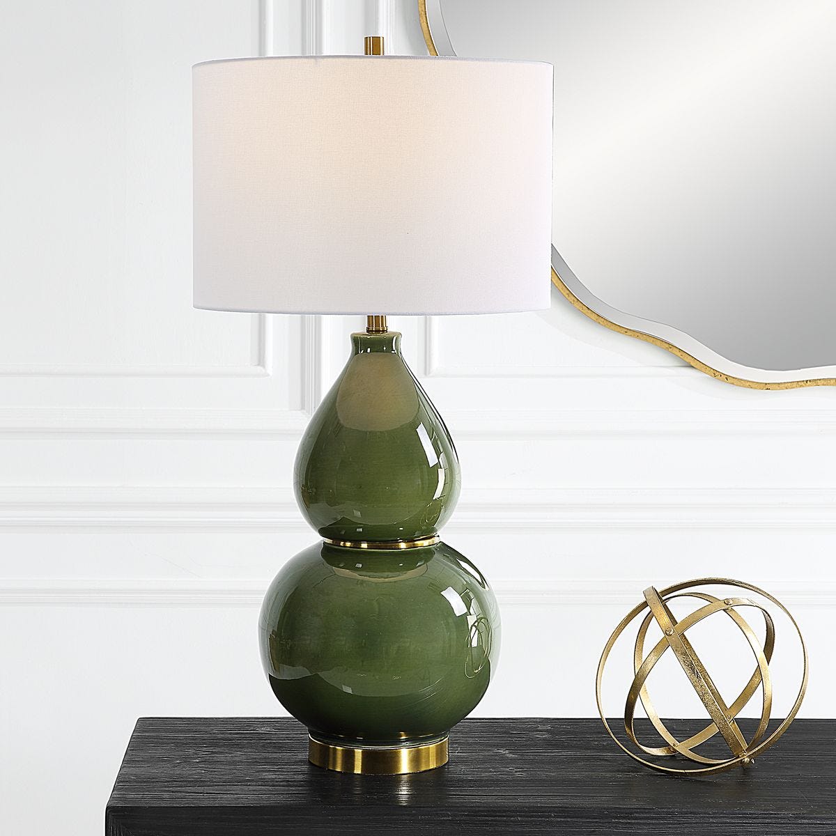 Green glass table lamp with white shade on a dark surface, with a gold decorative sphere and mirror in the background.