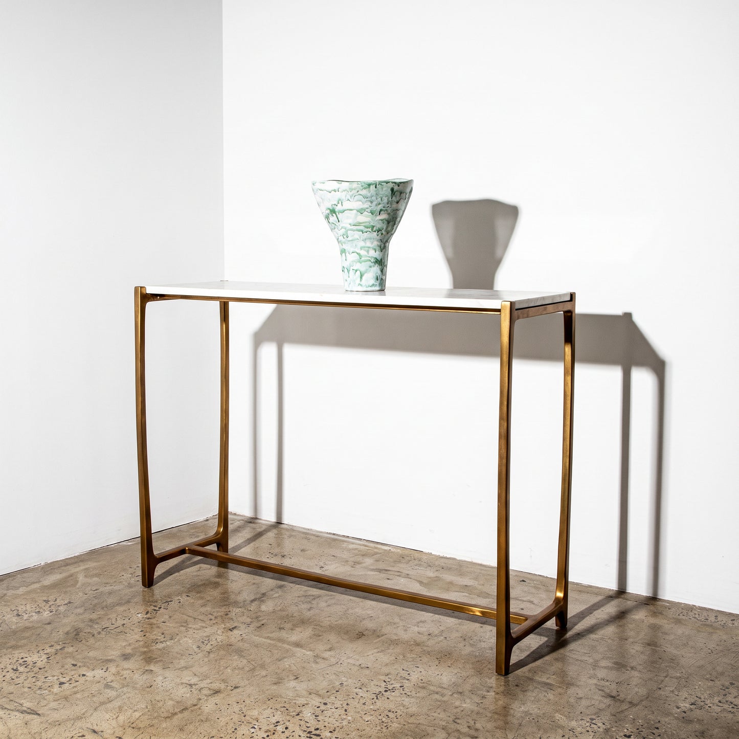 A modern console table with a white rectangular top and a slim gold metal frame, styled with a green and white marbled ceramic vase on a textured concrete floor against a white wall.