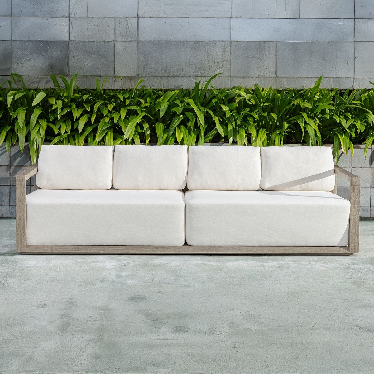 White outdoor sofa with wooden frame against a stone wall and green plants.