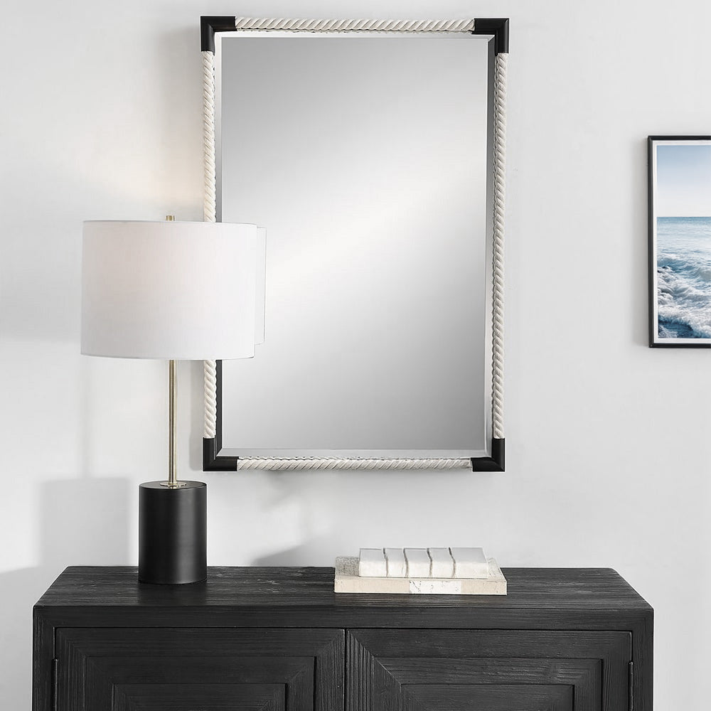 Nautical Mirror with White Rope frame and black corners over hanging a wood black console with a table lamp and white decorative books. An ocean photo hangs beside the mirror.