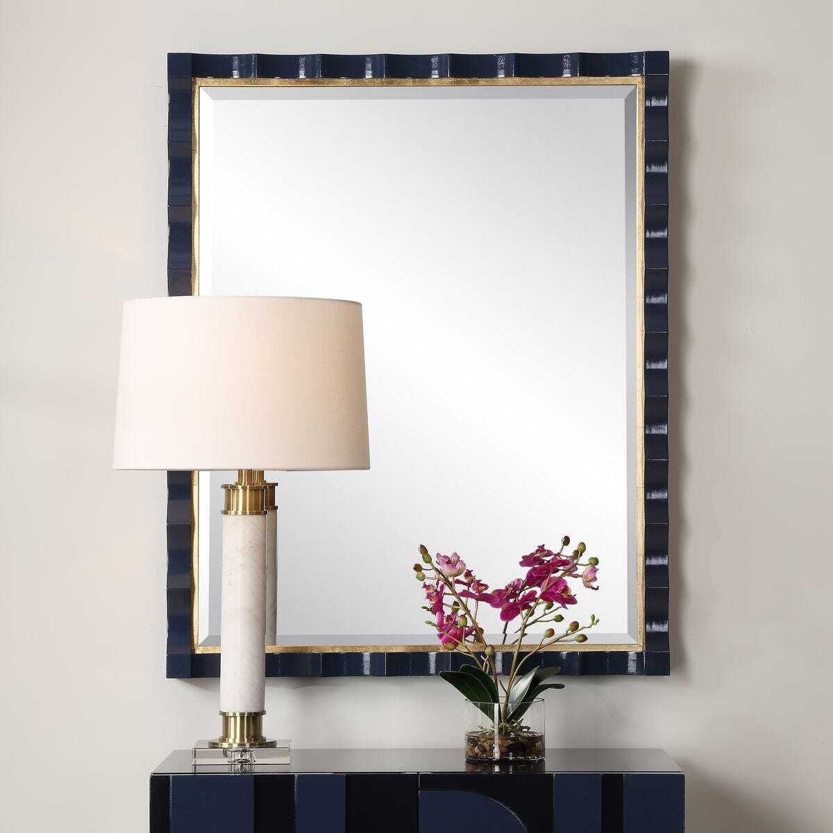 Decorative mirror with a lamp and flowers on a console table against a plain wall.