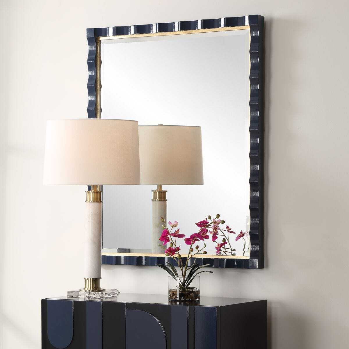 Decorative mirror with a black and gold frame above a dark blue console table with a lamp and flowers.