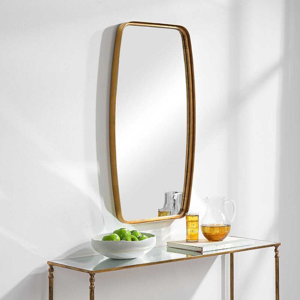 Gold-framed mirror on a white wall with a small table below holding a bowl of apples and glasses.