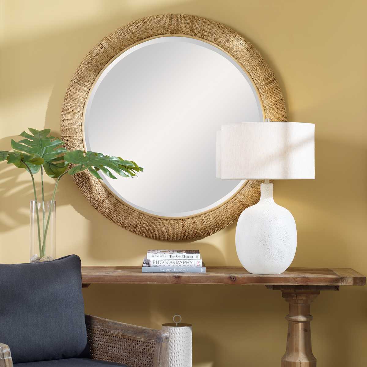 Round mirror with wicker frame above a wooden console table with a lamp and books against a yellow wall.