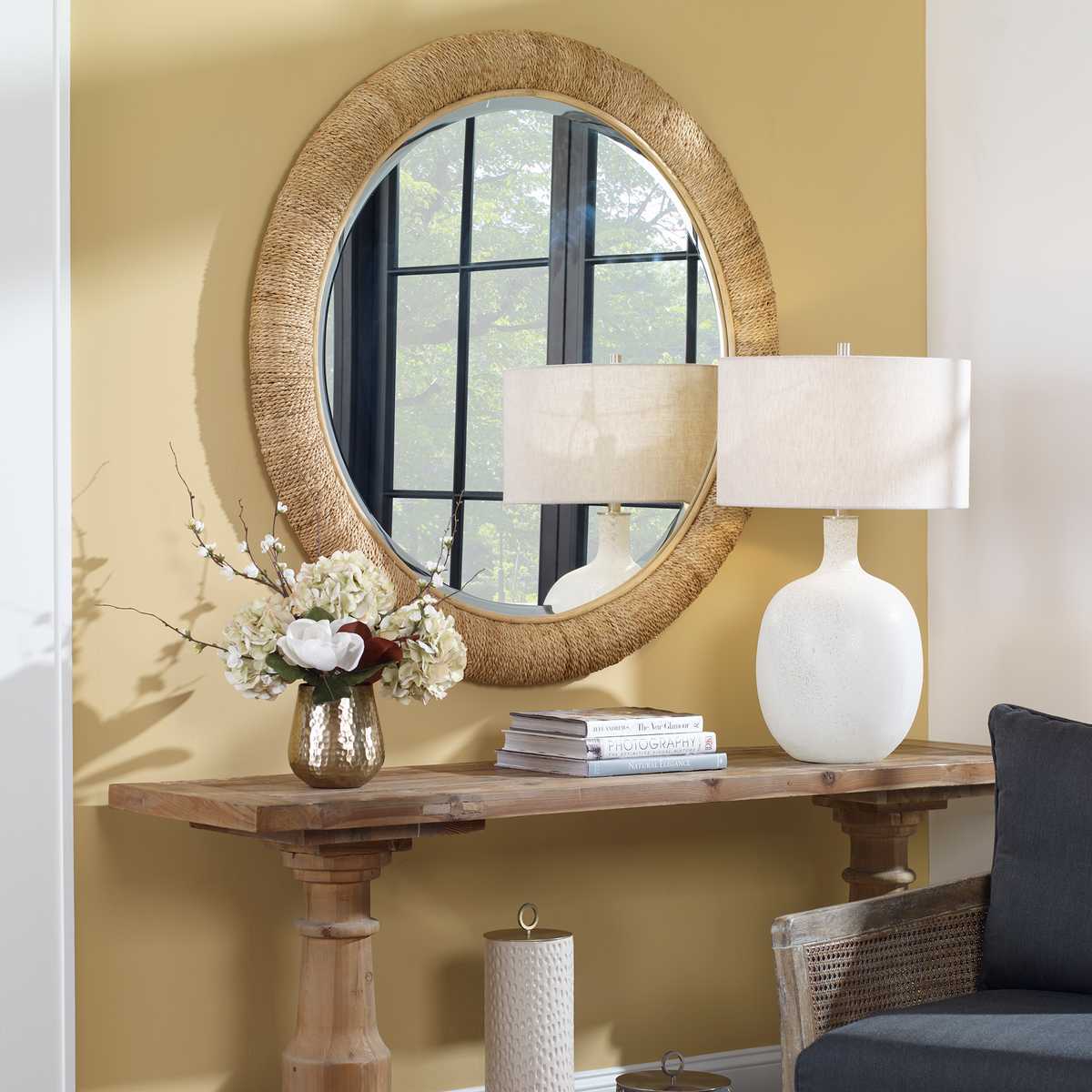 Round woven mirror on a wooden console table with lamps and decor items against a yellow wall.