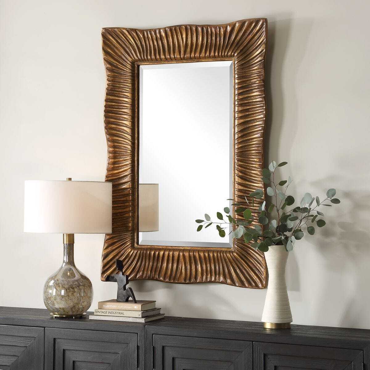 Decorative mirror with wavy frame on a dark wooden console table with lamp, books, and plant.