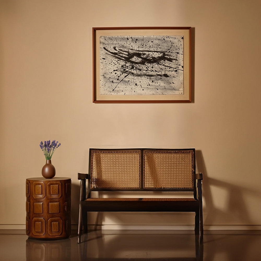Wooden bench with wicker backrest next to a carved wood side table with a vase, against a beige wall with a framed abstract painting.