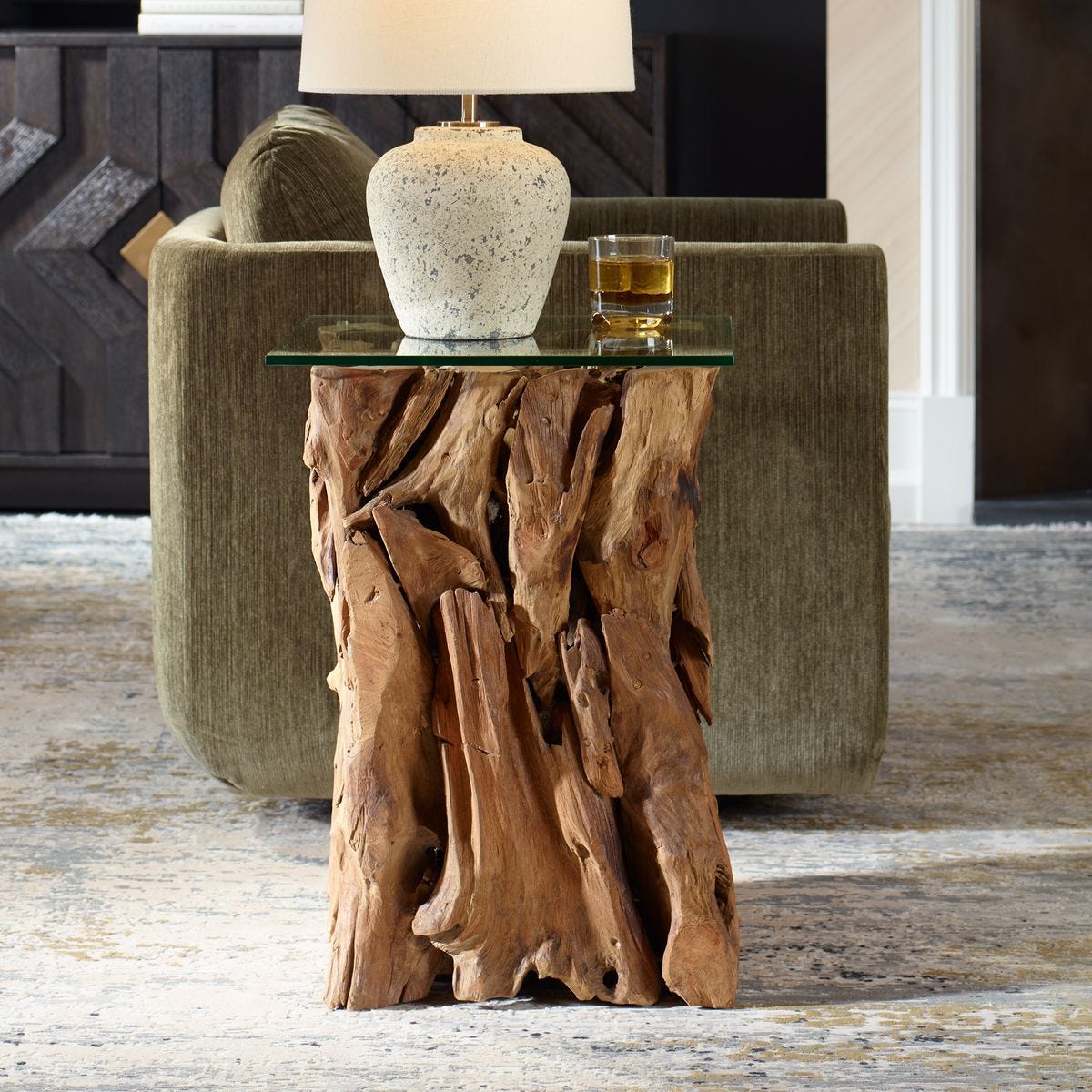 Wooden side table with a glass top holding a lamp and a glass of whiskey, in a living room setting.