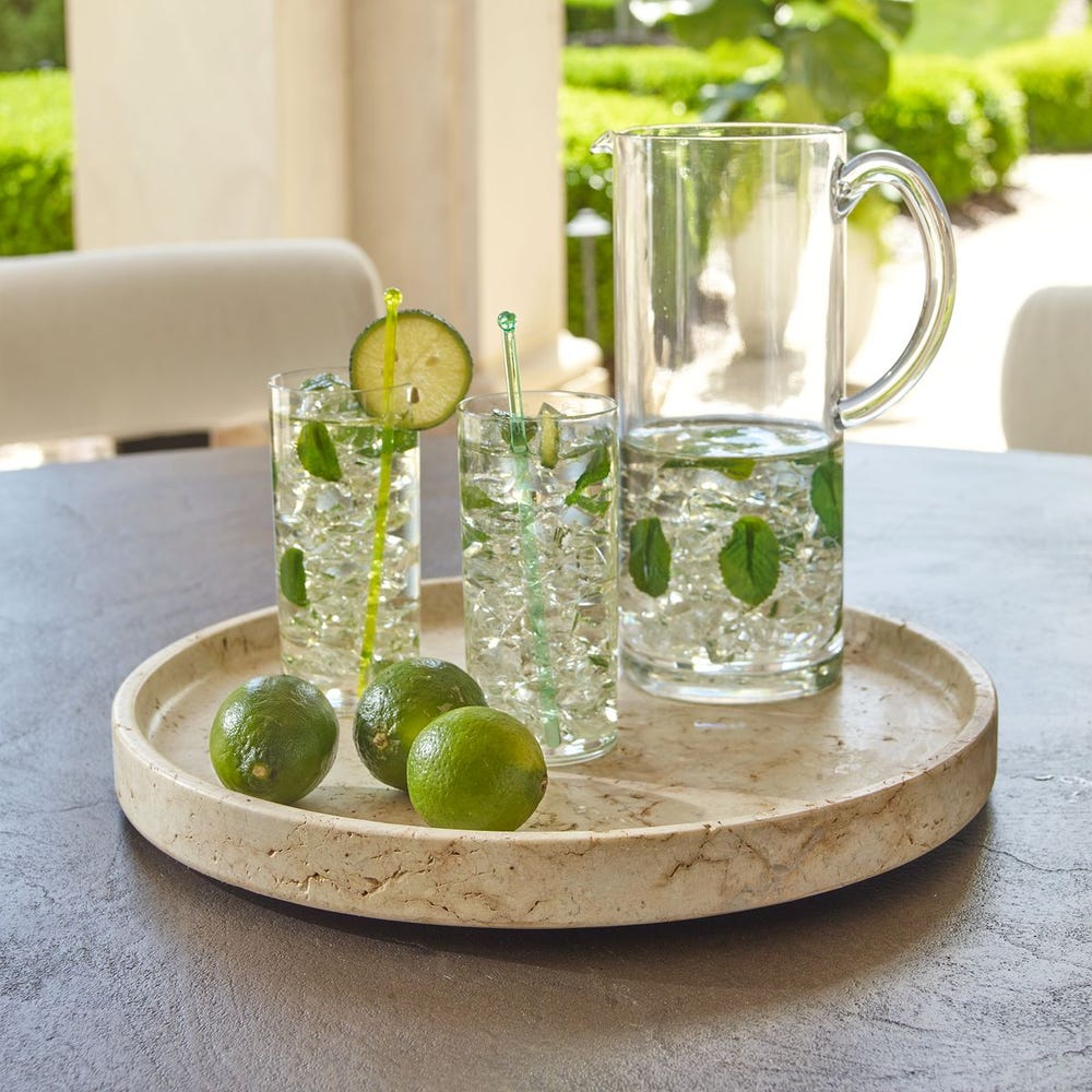 Clear glasses with lime slices and a pitcher on a travertine stone tray outdoors.