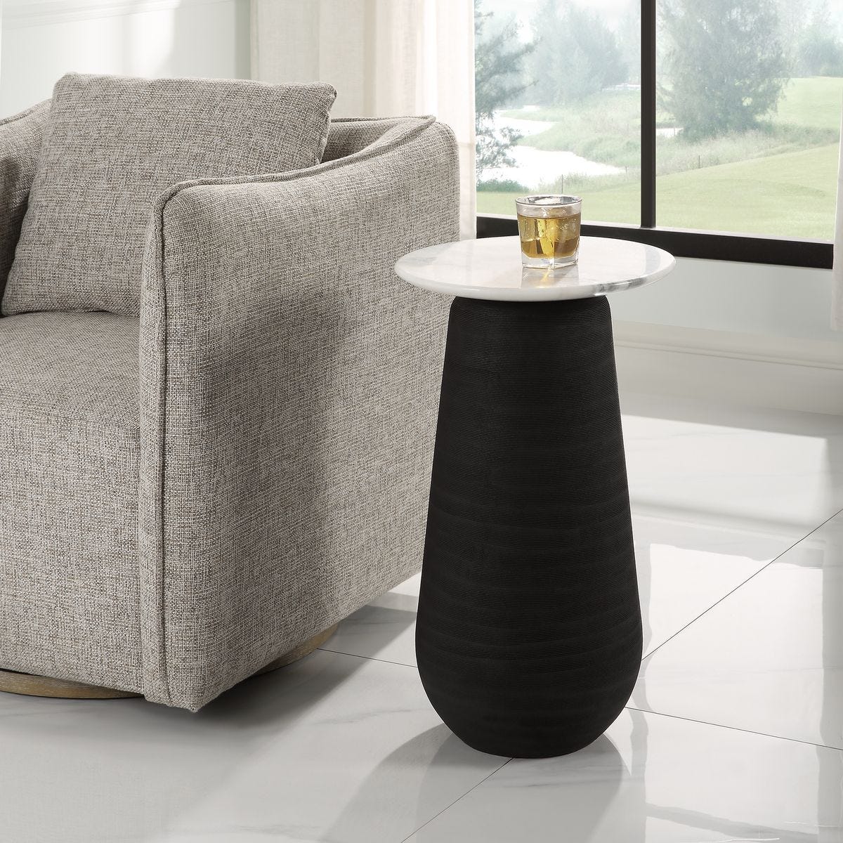 Black side table with a white round top next to a gray sofa in a room with large windows.