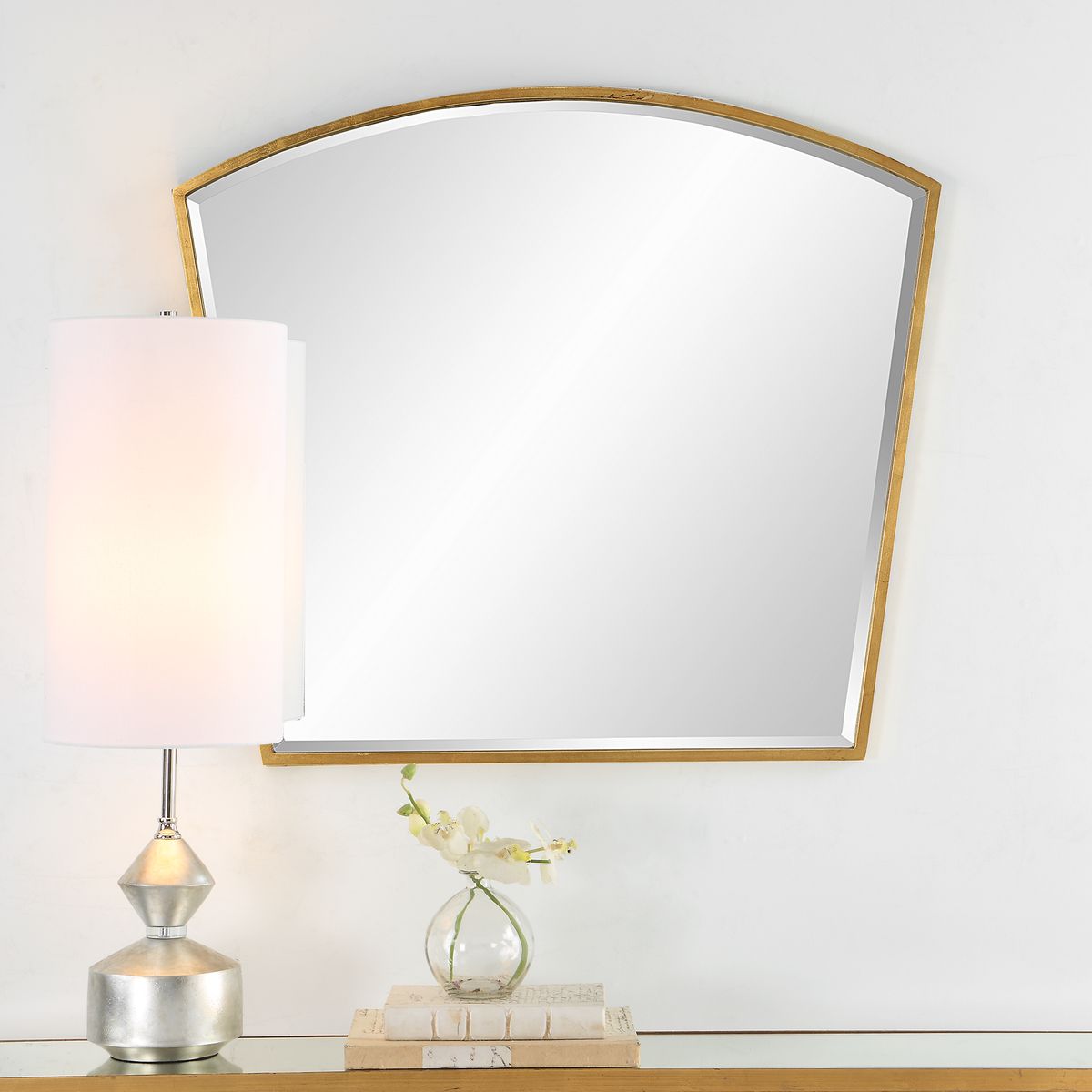 Decorative mirror with gold frame on a wall above a shelf with a lamp and small plant.