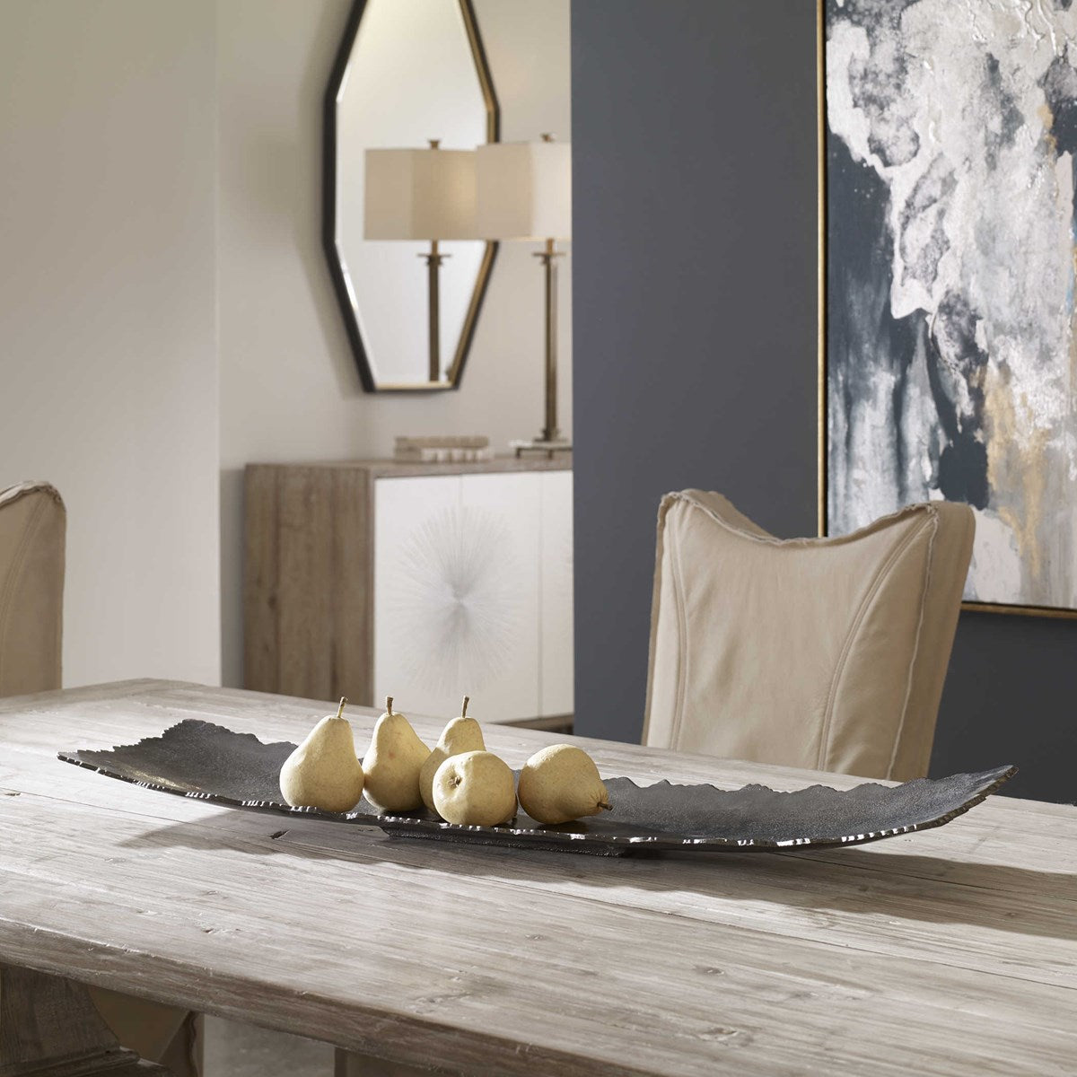 Nickel decorative tray on a modern, rustic wooden dining table with armchairs and an abstract painting in the background.