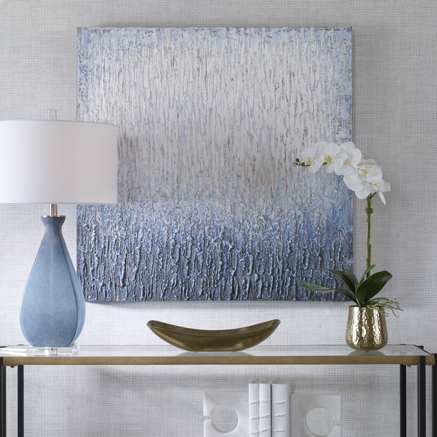 Hand painted piece of white and blue art behind a console with a lamp and white orchid flower.