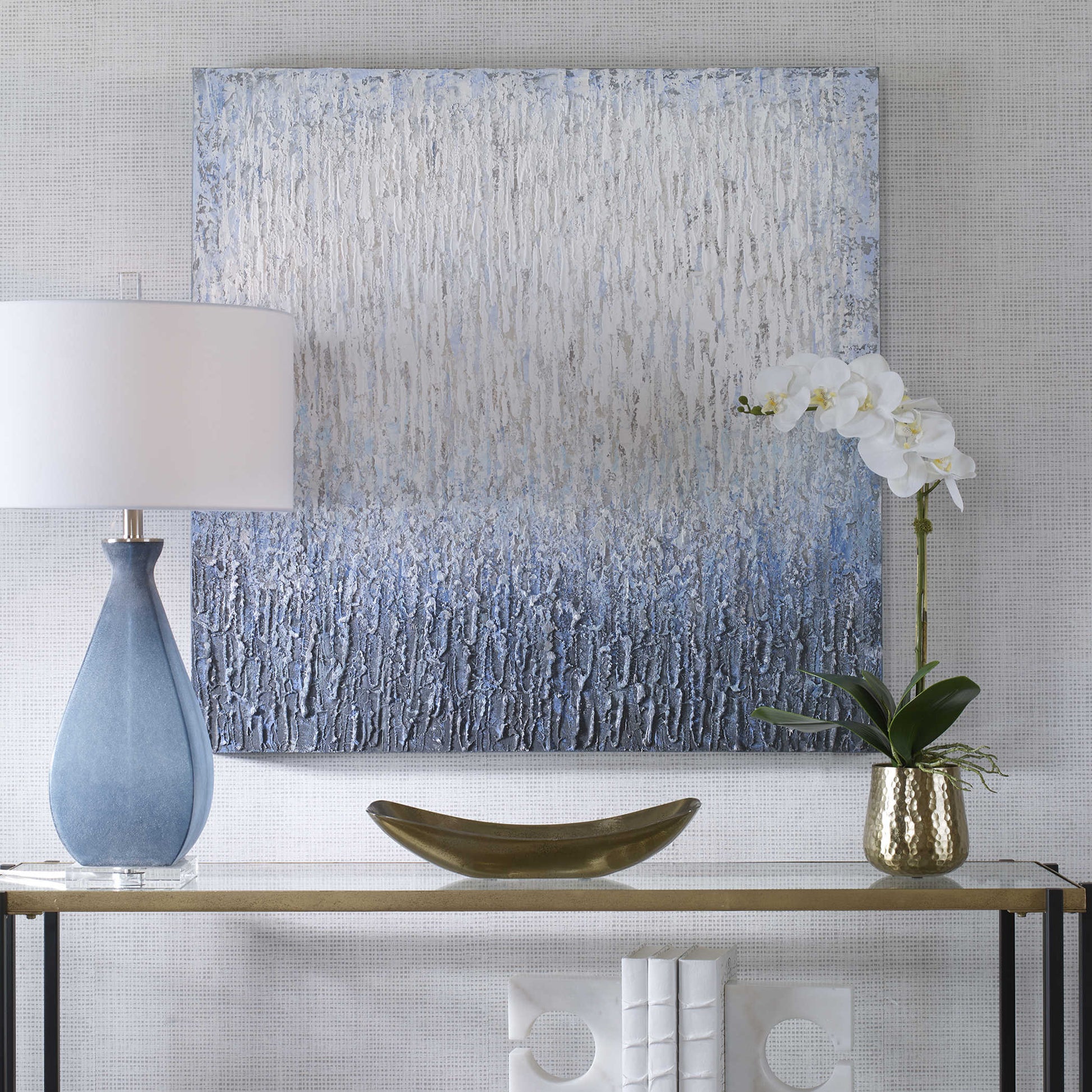 Hand painted piece of white and blue art behind a console with a lamp and white orchid flower.
