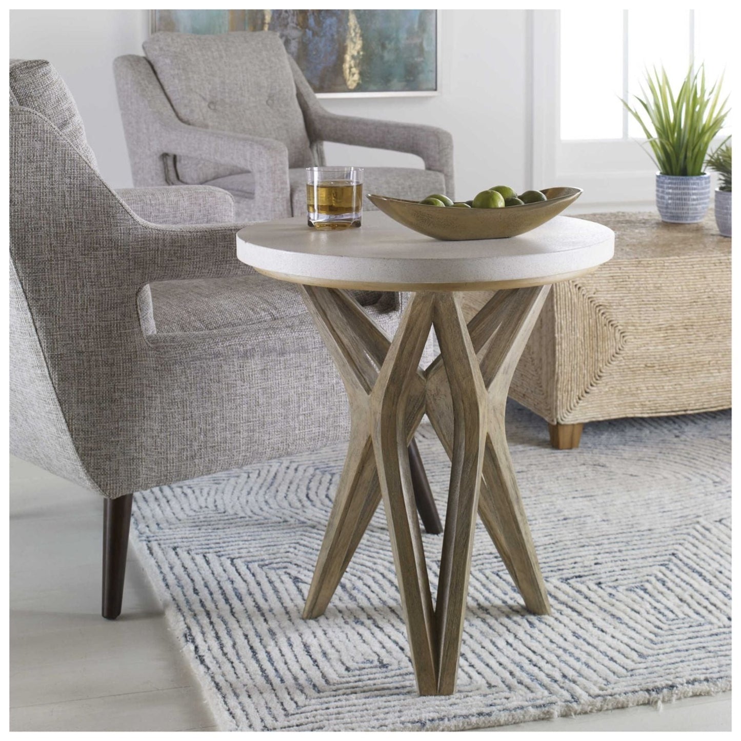 a natural ivory limestone top, on a geometric base finished in a warm oatmeal wash. Tsble showing bowl and a drink resting on top of table beside an occasional chair.