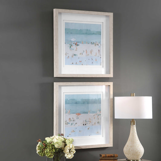 Stacked small art depicting a beach coastal scene on top of a console with a lamp, books, and flowers.
