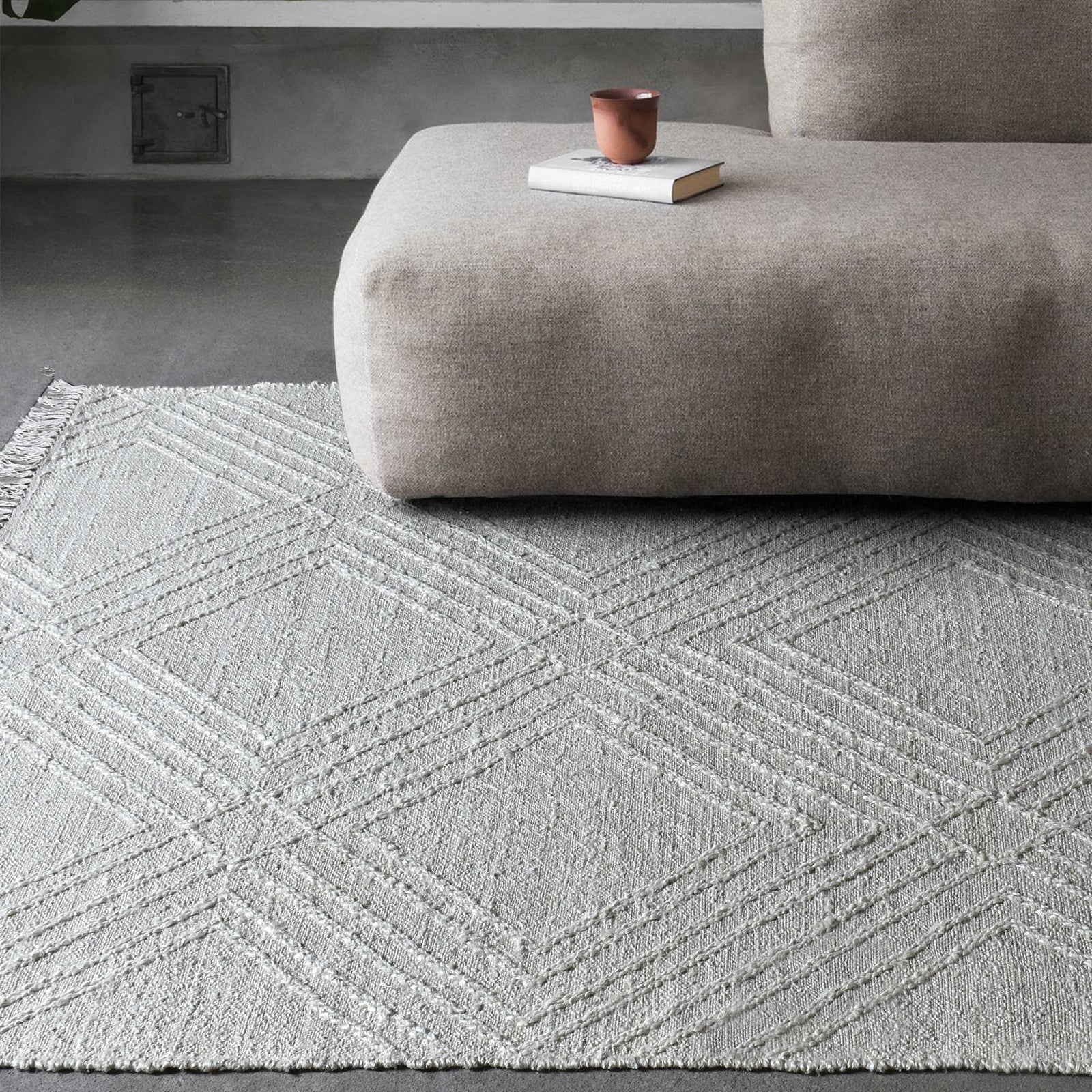 Geometric white rug on a concrete floor below a brown sofa with a book and cup on top.
