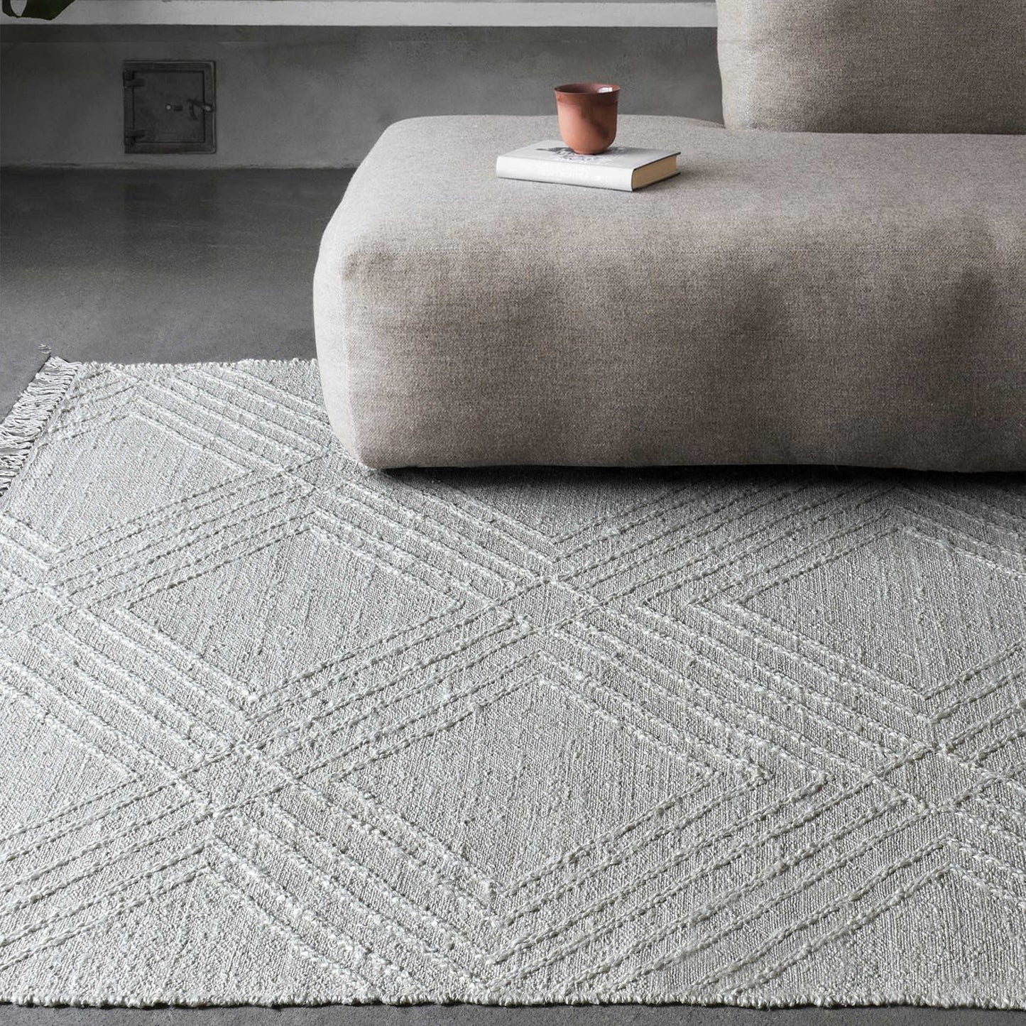 Geometric white rug on a concrete floor below a brown sofa with a book and cup on top.