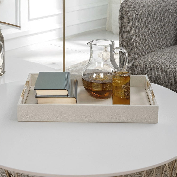 Coffee Table Tray with white faux leather finish and brass and acrylic handles decorated with books and a glass of iced tea on a white coffee table.