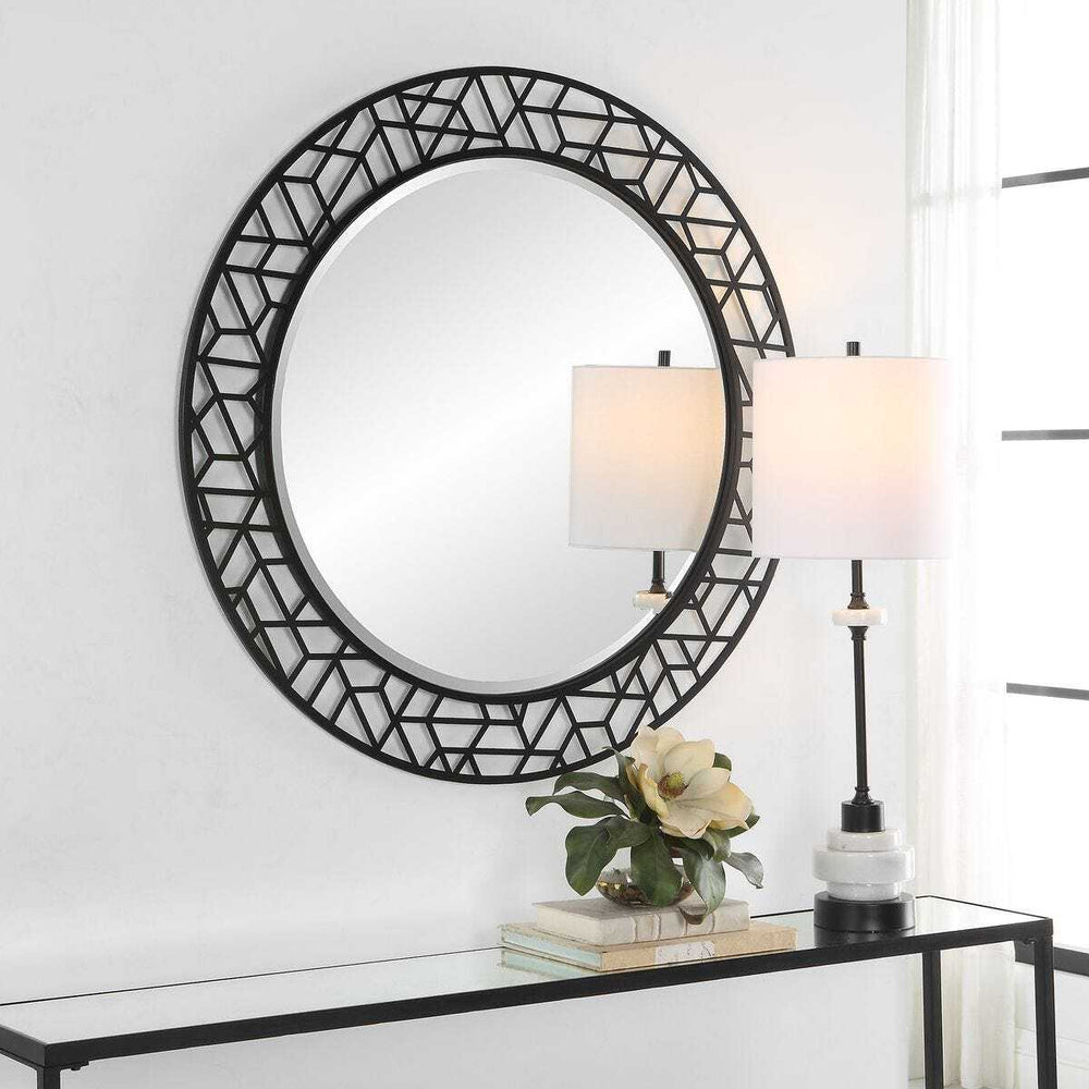 Round wall mirror with geometric black frame on a white wall, next to a table with decor items.
