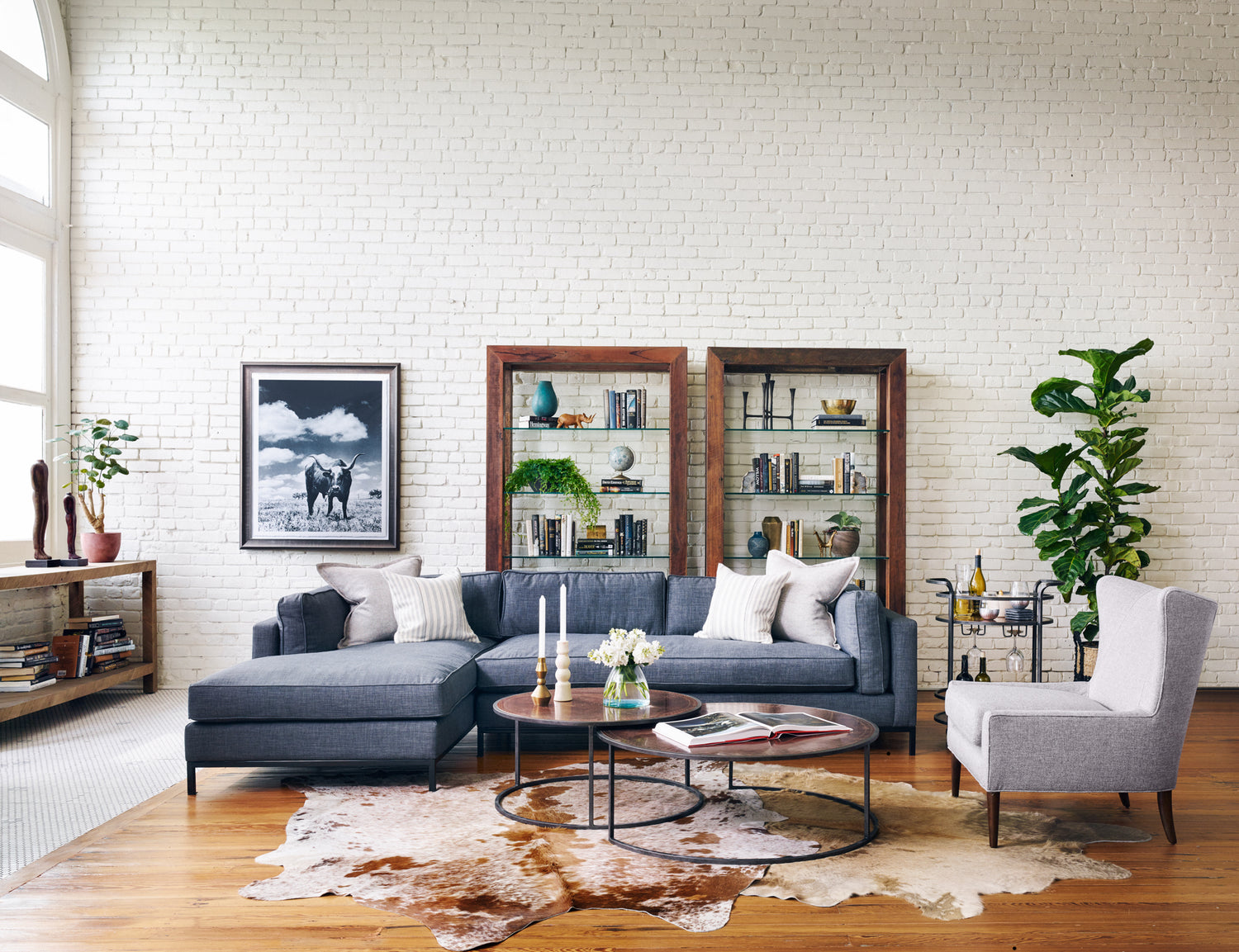 Expansive living room with bright light coming in on a blue couch with a round wood coffee table with white flowers and two book cases behind the couch. There is a grey armchair besides the coffee table with a fiddle leaf fig plan in the background. 