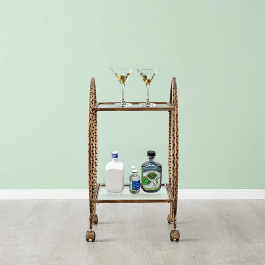 An old bart cart with hammered metal in the color antique gold. The back wall is a sea-foam green. Bottles of liquor are on the bottom shelf of the bar cart.