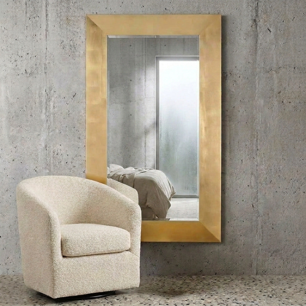 A large rectangular wall mirror with a thick brushed gold frame, leaning against a textured concrete wall next to a cream bouclé armchair. The mirror reflects a softly lit bedroom interior.