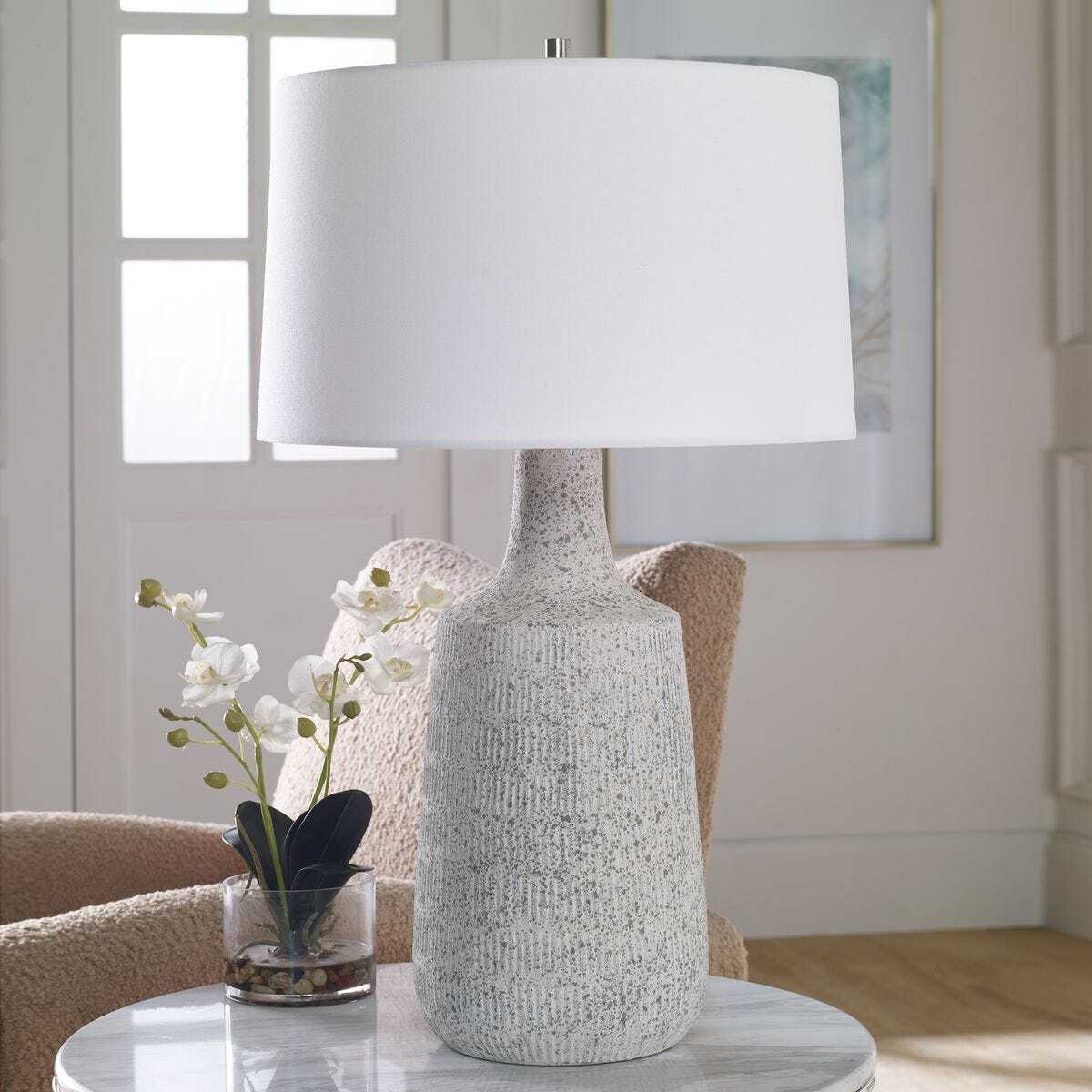 Table lamp with textured base and white shade on a marble surface in a room setting.