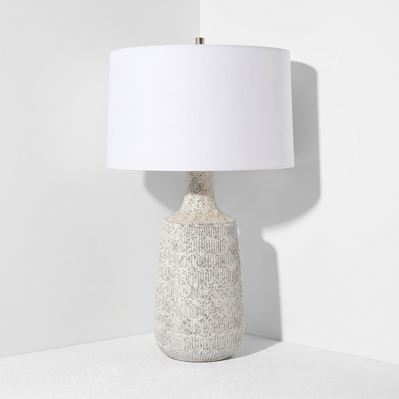 A table lamp with a textured, speckled off-white ceramic base and a large white drum shade, set against a clean white background.