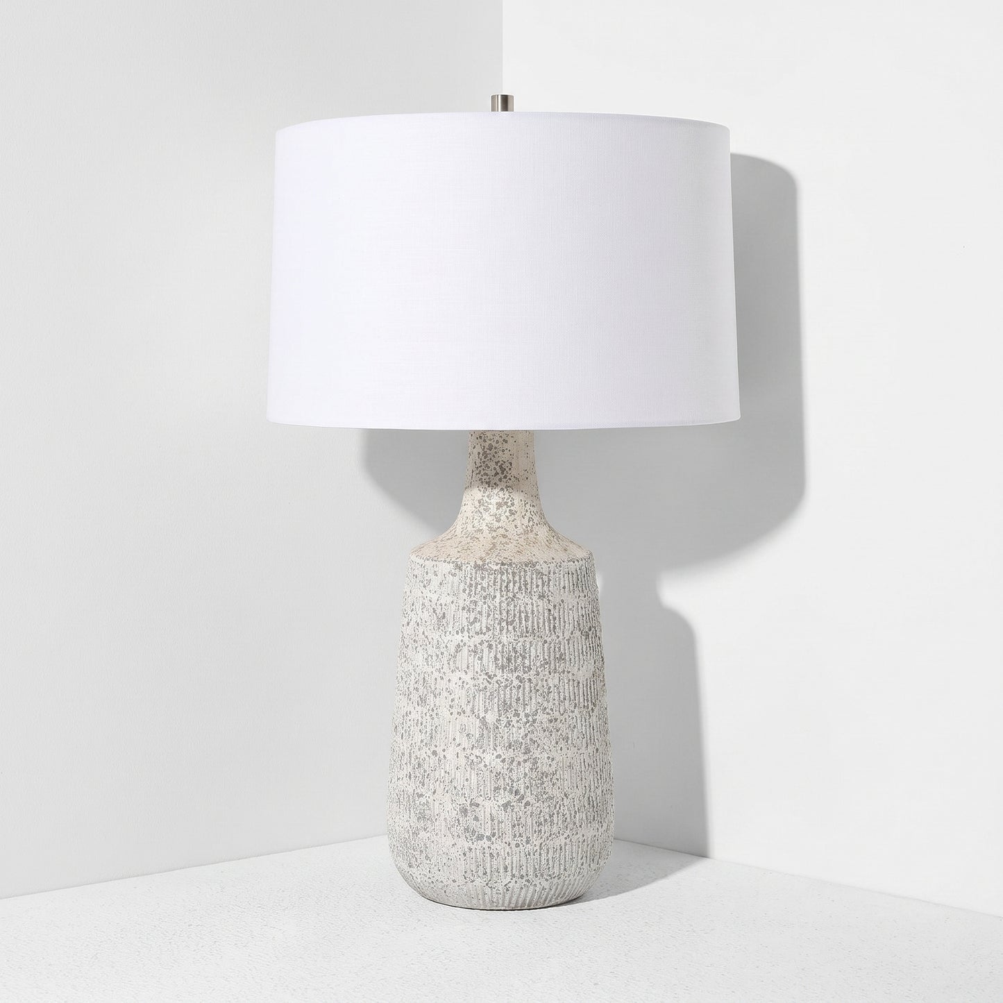 A table lamp with a textured, speckled off-white ceramic base and a large white drum shade, set against a clean white background.