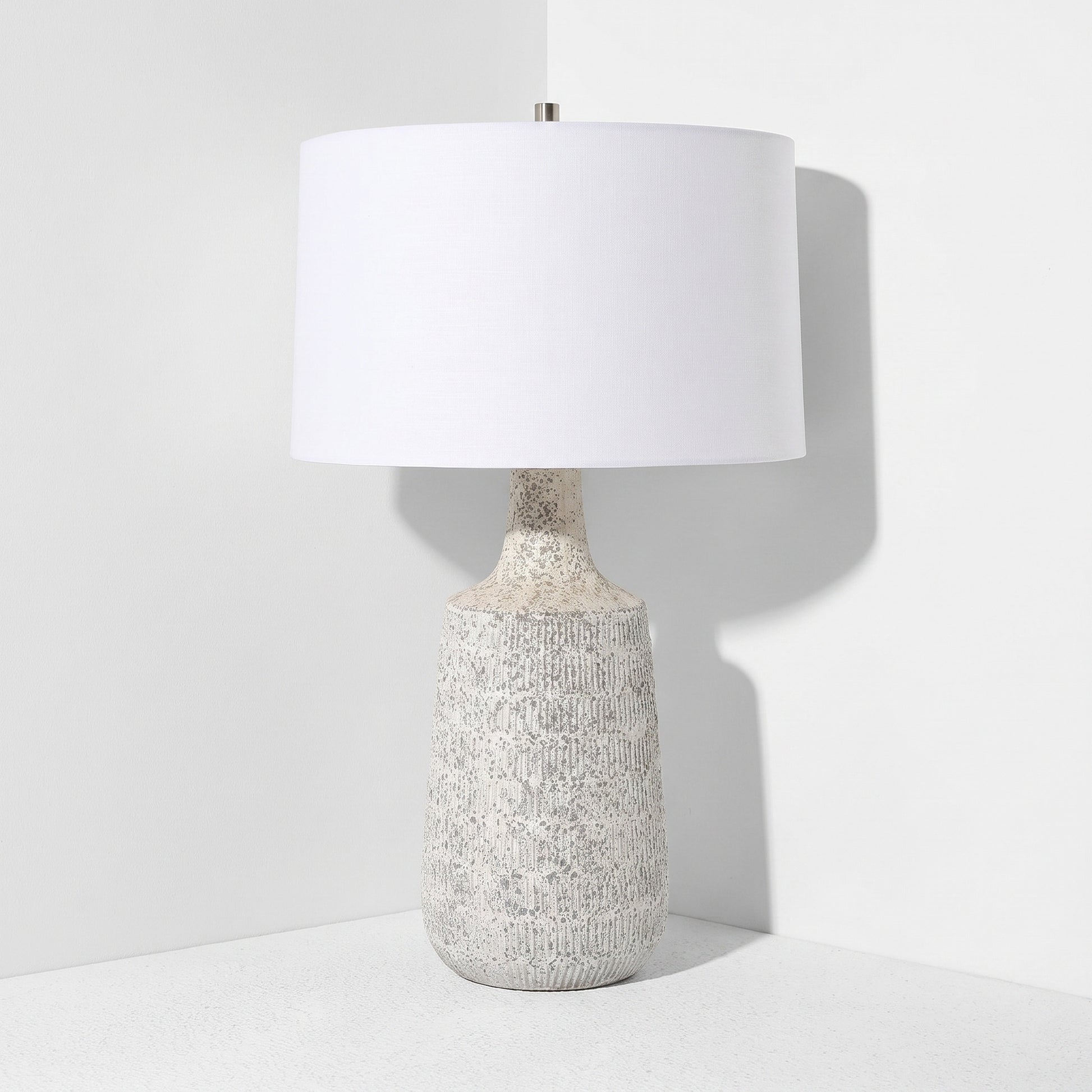 A table lamp with a textured, speckled off-white ceramic base and a large white drum shade, set against a clean white background.