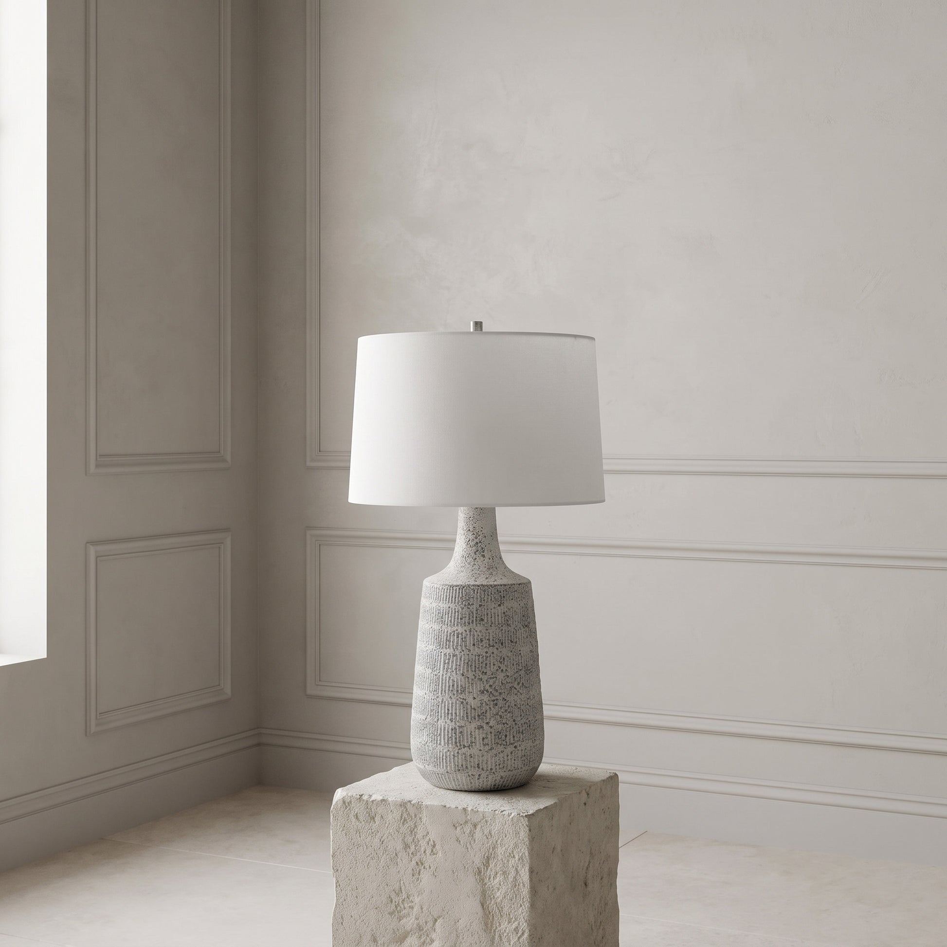 A table lamp with a textured gray ceramic base and a white drum shade, displayed on a stone block pedestal in a minimalist room with paneled walls.